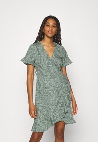 Vestido verde envolvente com bolinhas brancas, mangas curtas com babados, decote em V e barra com frufrus, feito de tecido leve com uma textura suave.