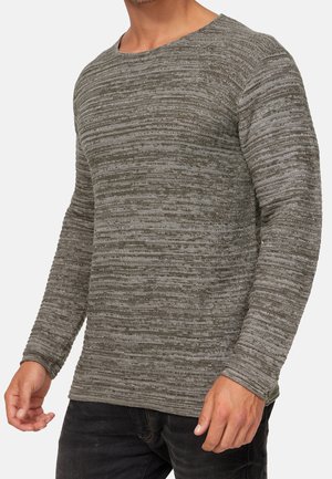 Man draagt een trui met lange mouwen en een ronde hals, met horizontale grijze en zwarte textuurranden, gecombineerd met een zwarte broek.