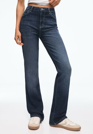 Vrouw draagt donkerblauwe rechte jeans en beige sneakers, staat met één hand bij haar taille.