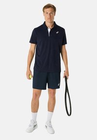 Marineblauw poloshirt met witte kraagdetail, gecombineerd met marineblauwe shorts. Accessoires omvatten een tennisracket en een groene tennisbal.