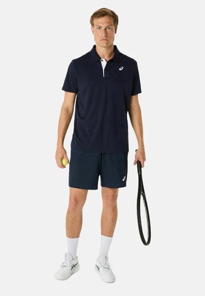 Polo bleu marine avec détail de col blanc, associé à un short bleu marine. Les accessoires comprennent une raquette de tennis et une balle de tennis verte.