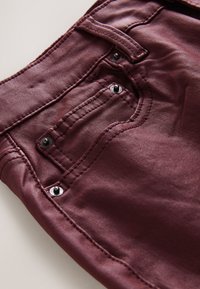 Burgundsko tkanino z gladko teksturo, ki ima izrazite šive in črne kovinske okraske na zadnjem žepu.