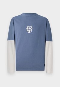 Blå langermet skjorte med rund hals, med et hvitt grafisk design på brystet. Ermene er kremfargede og laget av bomull.