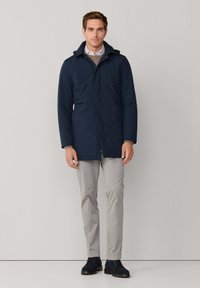 Dunkelblauer, wasserdichter Mantel mit Kapuze, Frontreißverschluss und seitlichen Taschen. Kombiniert mit hellgrauen Hosen und dunklen Schuhen. Schlichtes, funktionales Design.