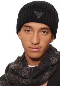 Bonnet côtelé noir avec un motif texturé, arborant un patch logo noir en forme de triangle. Accompagné d'une écharpe noire à motif.