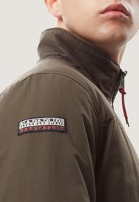 Oliven grøn jakke med høj krave, der har lynlås lukning og rød snor. Broderet logo patch på venstre skulder. Glat tekstur.