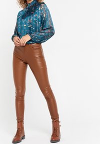 Teal blommig genomskinlig blus med hög krage, parat med figurnära bruna fusk läderbyxor och bruna snörda anklarstövlar med spännardetaljer.