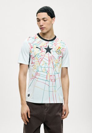 Fiatal férfi fehér Ghánai nemzeti labdarúgócsapat mezt visel, színes geometriai vonalakkal, fekete csillaggal és csapatemblémával.