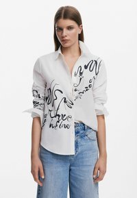 Camicia bianca con bottone e design di testo artistico nero sul davanti. Presenta maniche lunghe e una vestibilità ampia, abbinata a jeans in denim azzurro chiaro.