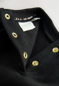 Polo nera con colletto morbido, dettagli in metallo color oro, cuciture visibili e etichetta bianca con testo verde.