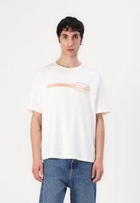 White cotton t-shirt with short sleeves and a round neckline. Features a colorful "diesel" graphic across the front. Worn with blue jeans.
