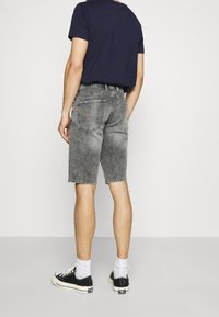 Mies, który ma na sobie przetarte czarne dżinsowe szorty, granatowy t-shirt, białe skarpetki i czarne niskie trampki, stoi tyłem na jednolitym tle.