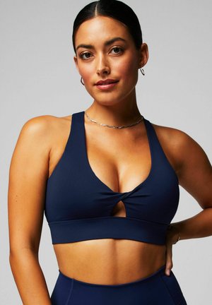 Soutien-gorge de sport bleu marine avec un décolleté plongeant et un design à l'avant torsadé. Il présente une large bande et un tissu lisse et extensible.