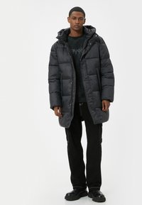 Puffjakke i mørkegrå med quiltet design og hætte, med frontlynlås og trykknaplukninger, parret med sorte bukser og støvler.