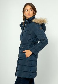 Námorně modrá péřová bunda se zipem vpředu, umělou kožešinou na kapuci, elastickým pasem a bočními kapsami. Má prošívanou texturu a kovové akcenty.