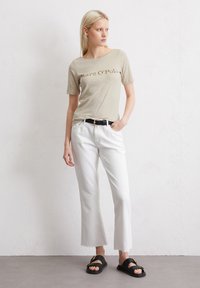 Camiseta de manga corta beige con el logo "Marc O'Polo", combinada con pantalones de pierna ancha blancos y sandalias planas negras sobre un fondo neutro.