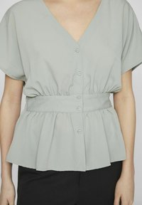 Gaiši pelēka, īsām piedurknēm blūze ar V veida kakla izgriezumu un pogu aizdari, ar elastīgu jostasvietu pepluma formai un mīkstu, gludu tekstūru.