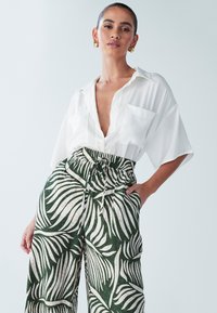 Bela majica kratkih rokavov z globokim V-izrezom in dvema žepoma na prsih, kombinirana z zelenimi lanenimi hlačami z belim vzorcem palmovega lista.