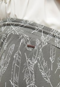 Šedá látka s potiskem bílých peříček, vybavená elastickým pasem a malou hnědou etiketou s nápisem "noppiess" připevněnou na straně.