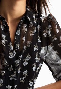 Blusa nera trasparente con maniche corte, motivo floreale in bianco e verde, dettaglio del colletto e chiusura con bottoni.