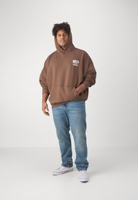 Hanorac oversized maro cu buzunar frontal, detaliu grafic alb, asociat cu blugi din denim de un albastru deschis și teniși albi. Design fără cusături și textură moale.