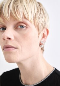 Pendientes de plata con un diseño circular texturizado y un acento geométrico, llevados con el pelo corto. Top negro con un escote de cuentas.