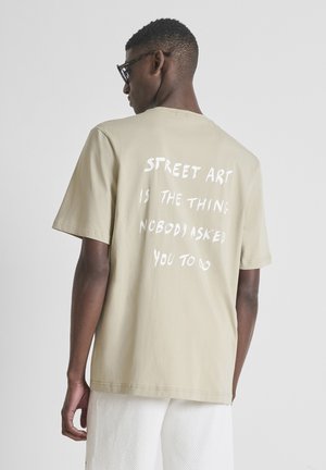Muškarac u bež majici s bijelim tekstom "STREET ART IS THE THING NOBODY ASKED YOU TO DO" i bijelim teksturiranim kratkim hlačama, okrenut leđima.