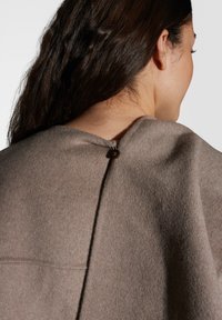 Bruine wollen kleding met een ronde kraag en een decoratieve knoopsluiting aan de achterkant, met een gladde, zachte textuur.