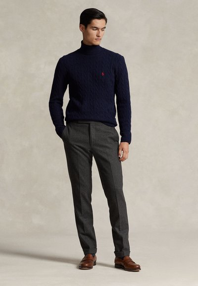 Uomo in piedi con le mani in tasca, indossa un maglione dolcevita blu navy, pantaloni grigi e scarpe di cuoio marroni su sfondo uniforme.