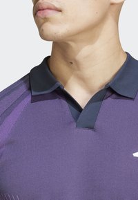 Polo de color púrpura con un acabado texturizado, cuello en contraste en un oscuro tono de navy, y diseño de escote en V. Patrones diagonales sutiles en las mangas.