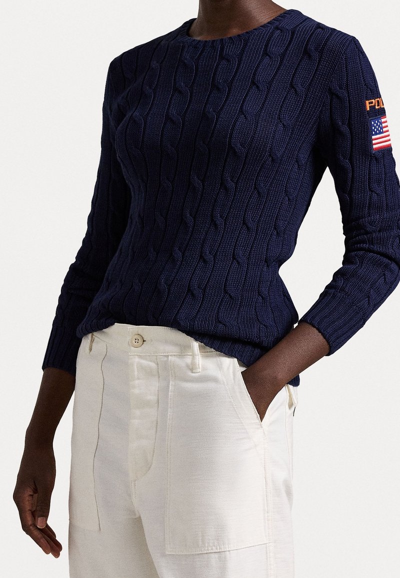 Maglione blu navy a trecce con maniche lunghe, scollo rotondo e toppa con la bandiera americana sul braccio sinistro, abbinato a pantaloni color crema.