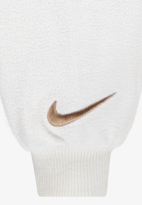 Balta aplokļa džeza jaka ar ribotu piedurkni un bronzas izšūtu Nike swoosh logo. Mīksta, teksturēta virsma bez redzamām rakstāmzīmēm.