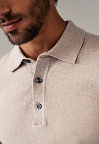 Beige stickad polo med en mjuk textur, som har en krage och tre mörka knappar framtill.
