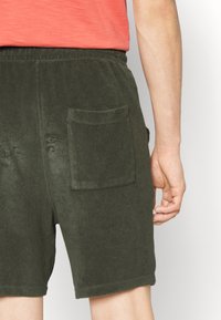 Shorts en éponge verte avec une poche arrière plaquée, une taille élastique et une texture lisse. Coutures visibles et détail de l'ourlet.