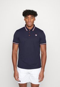 Marine blauwe polo shirt van een katoenmix, met rode en witte gestreepte kraag en mouwen, voorzien van een logo op de linkerkant van de borst en knopen.