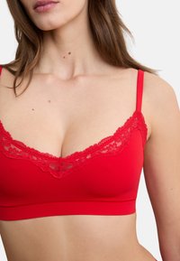 Frau trägt ein rotes Bralette mit Spitzenbesatz und dünnen Trägern, zeigt Oberkörper und Hals vor weißem Hintergrund.