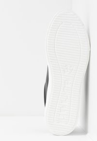 Chaussure noire avec une semelle en caoutchouc blanche, présentant un motif de grip texturé. Le talon est légèrement surélevé et lisse, avec un design minimaliste.