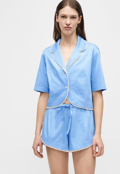 Γυναίκα φορώντας ανοιχτό γαλάζιο κοντομάνικο πουκάμισο με κουμπιά και ταιριαστά σορτς με κορδόνι και λευκή επένδυση, στέκεται μπροστά σε απλό φόντο.