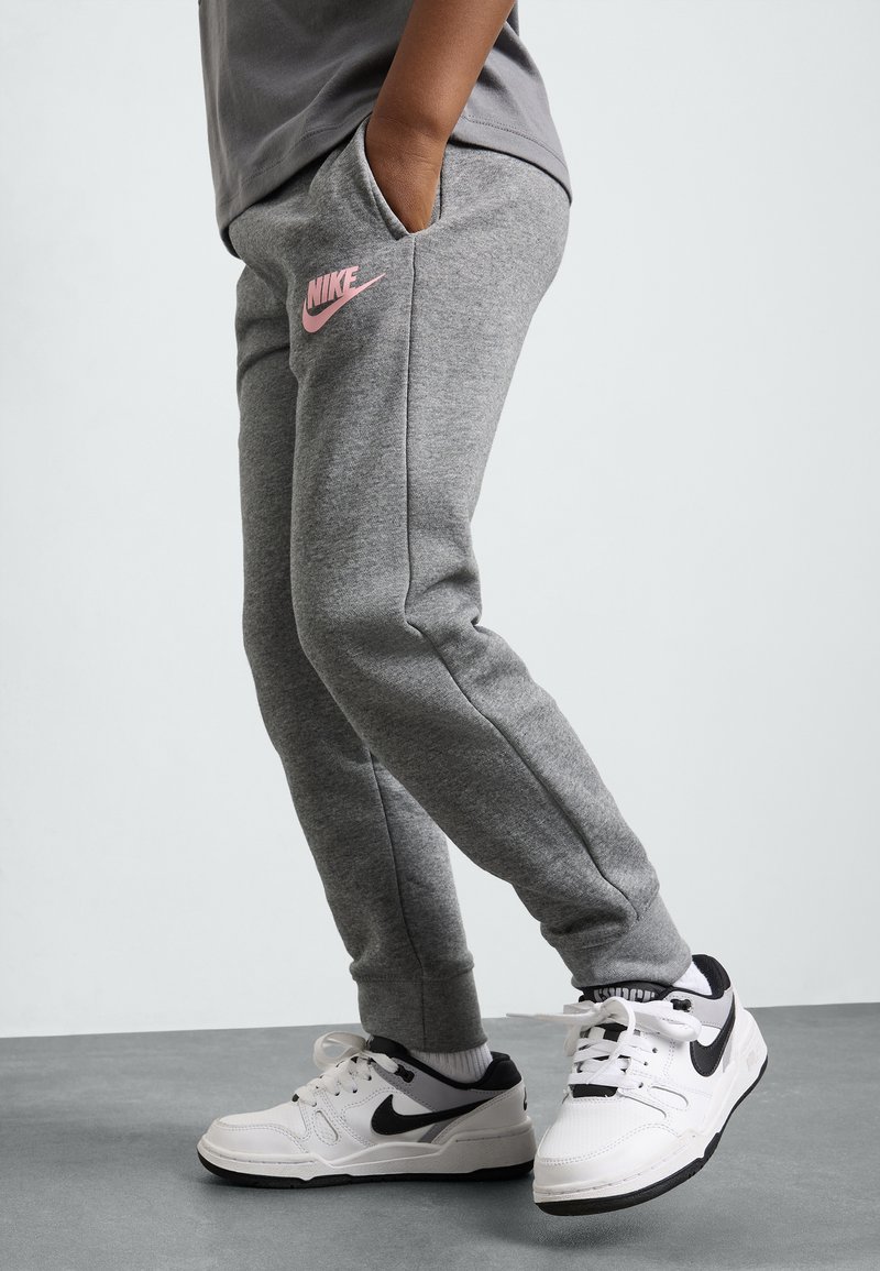 Calças de moletom cinzentas com um logo da Nike rosa, com corte ajustado e bolsos, combinadas com ténis Nike brancos e pretos sobre uma superfície cinzenta.