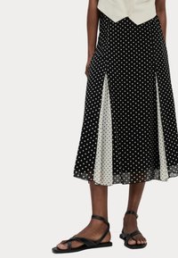 Robe midi à pois noirs et blancs avec des panneaux transparents contrastants, présentant un design évasé. Portée avec des sandales plates noires.