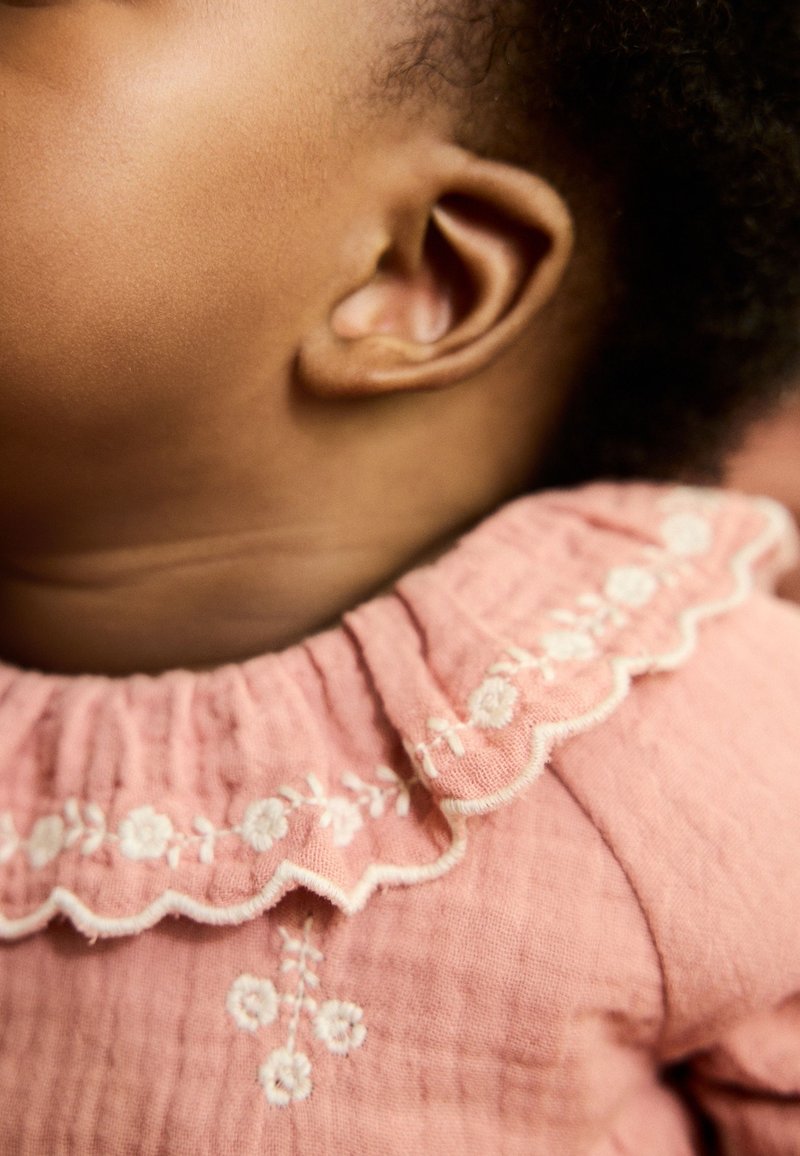 Vêtement en coton rose avec un col festonné et une broderie florale blanche. Tissu texturé et gros plan sur une petite oreille.