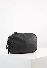 Černá kožená crossbody taška obdélníkového tvaru s horním zipovým uzávěrem a dekorativními třásněmi na jedné straně. Hladký povrch.