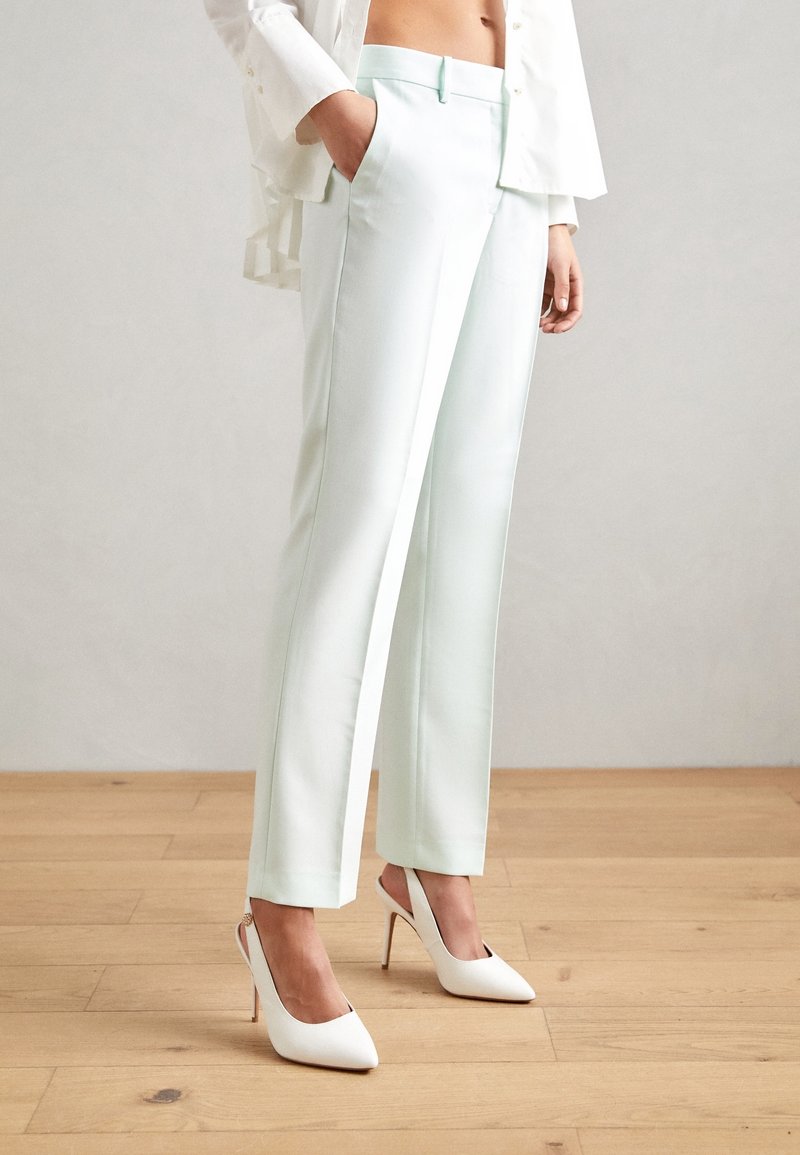 Femme portant un pantalon blanc sur mesure et des chaussures blanches à talons hauts, debout sur un sol en bois avec la main dans la poche.