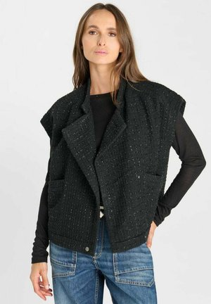 Frau trägt eine schwarze, strukturierte ärmellose Weste über einem schwarzen Langarmshirt und blauen Jeans, steht mit einer Hand in der Hüfte.