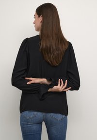 Juoda ilgaa rankovė bluzonė su pūstais pečiais ir raukšlėtais rankogaliais, derinama su mėlynais džinsais, matoma iš nugaros.