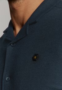Close-up van een persoon die een donkerblauwe button-up shirt draagt met een klein zwart en geel hexagonaal logo op de borst.