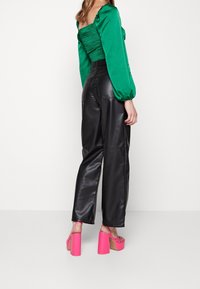 Zelený top s nabíranými rukávy a smockovaným detailem, zkombinovaný s vysokými černými kalhotami z faux kůže a růžovými platformovými podpatky.