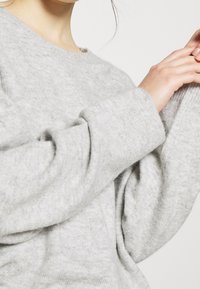Donna che sistema la manica del suo maglione morbido e grigio chiaro, con maniche larghe e leggermente oversize, su uno sfondo semplice.