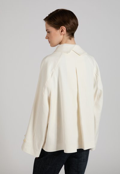 Donna con capelli corti castani indossa una giacca crema oversize con dettaglio plissettato sulla schiena e maniche larghe, abbinata a pantaloni scuri.