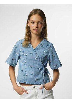 Jeune femme portant un top portefeuille en denim bleu clair avec de petites fleurs brodées et un pantalon blanc, les mains dans les poches.